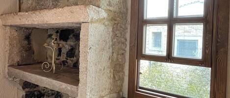 A stone fireplace with a wooden shelf, a window with a view of greenery, and a patterned rug on the floor.