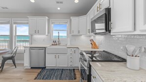 A modern kitchen with white cabinets, a stainless steel stove, and a view of the ocean through the windows. - Beachfront, HotTub, Elevator, Hammocks, Pet & Familly Friendly (Surf City)