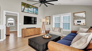 A modern living room with a large flat-screen TV, a ceiling fan, a coffee table, a sofa, and a dining area with a wooden cabinet. - Beachfront, HotTub, Elevator, Hammocks, Pet & Familly Friendly (Surf City)