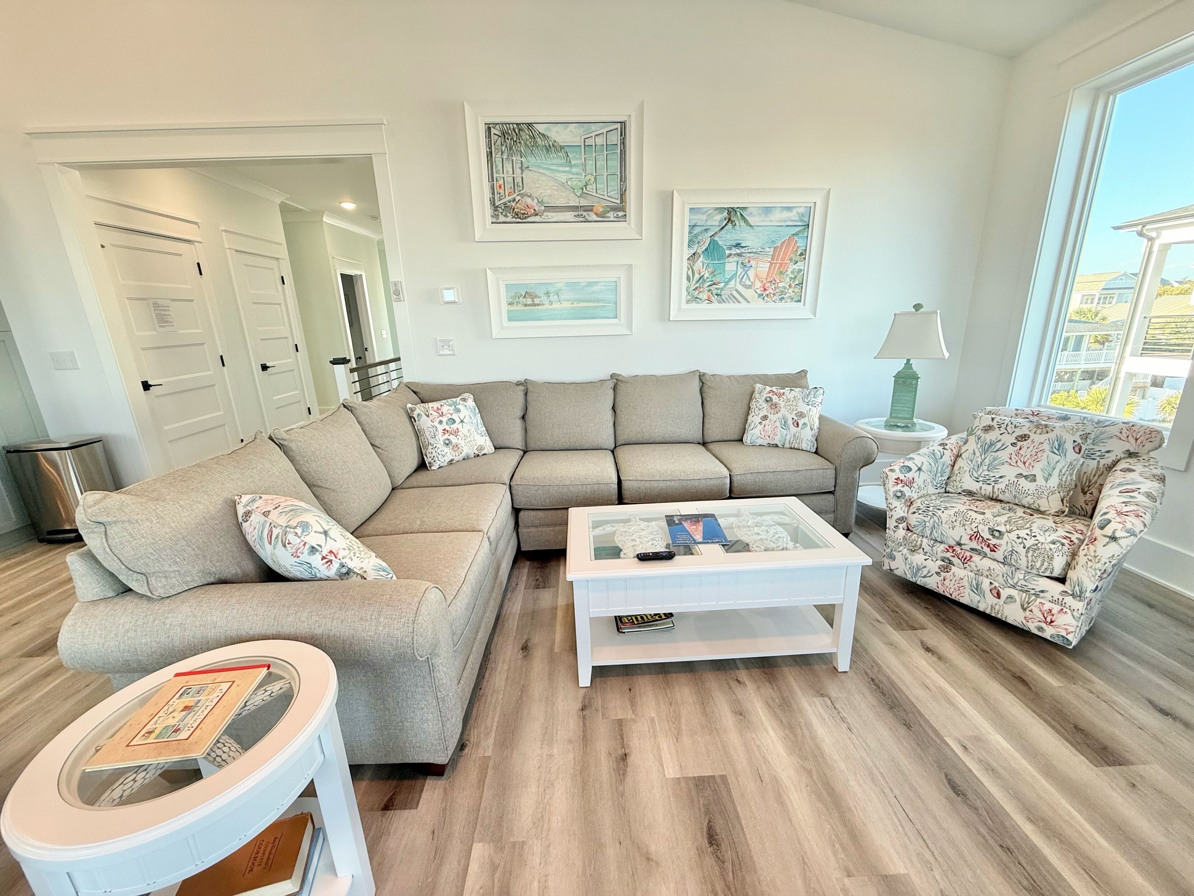 A modern living room with a beige sectional sofa, a white coffee table, and a floral armchair. There are framed artworks on the wall and a view of the outdoors through a large window.