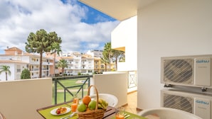 A balcony with a table set for breakfast, including fruits and juice, overlooking a residential area with white buildings and palm trees.