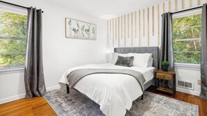 A bedroom with a bed, a bedside table, a wall-mounted shelf, and a window with curtains.