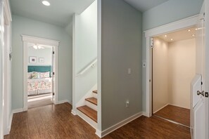 Um corredor com piso de madeira, escada e porta que dá para um quarto com cama e janela.