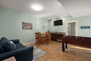 Ett vardagsrum med en mörkblå soffa, ett matbord i trä med stolar, ett fotbollsspel, en köksdel med en tv och en skylt med texten ”Avkoppling” på väggen.