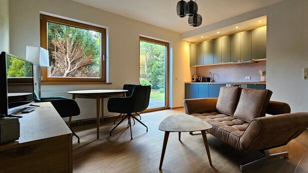 Ein modernes Wohnzimmer mit Sofa, Esstisch und Stühlen. Es gibt eine Küchenzeile mit Schränken und einem Fenster.