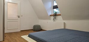 Une chambre avec un plafond en pente, un lit, un radiateur, une porte et un petit tabouret.