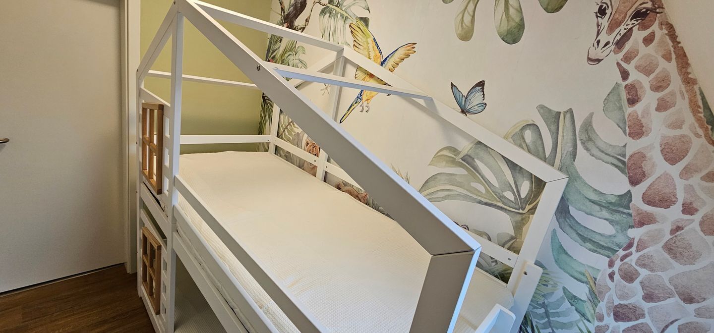 Een kinderbed met twee bedden, met een wit frame en een matras, voor een muur met een giraf- en vlindermuurdecoratie.
