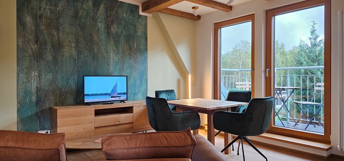 Un salon moderne avec un meuble TV en bois, un canapé et une table à manger avec des chaises. Une grande fenêtre offre une vue sur l’extérieur.