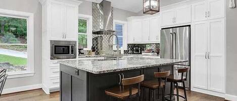 Una cocina con armarios blancos, una isla grande con taburetes de bar, un refrigerador de acero inoxidable y una lámpara colgante.