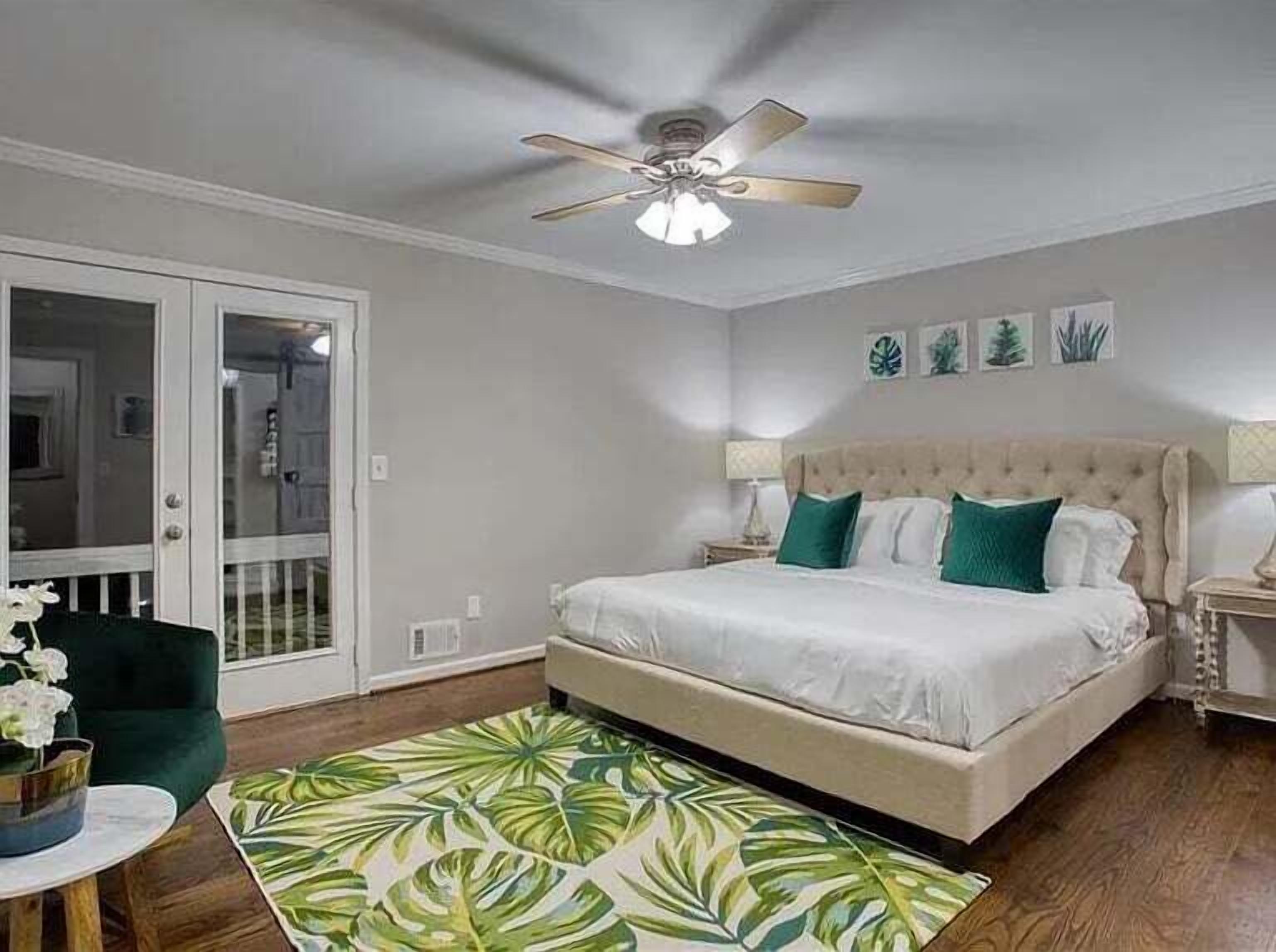 Une chambre avec un grand lit, un ventilateur de plafond, une table de chevet, un fauteuil vert et une vue sur l’extérieur grâce à une porte-fenêtre coulissante.