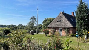 A thatched-roof house with a brick chimney, surrounded by a well-maintained garden with various plants and flowers. - Newly renovated vacation home - 4 min. to the beach - large, quiet lot surrounded by nature  (Schashagen)