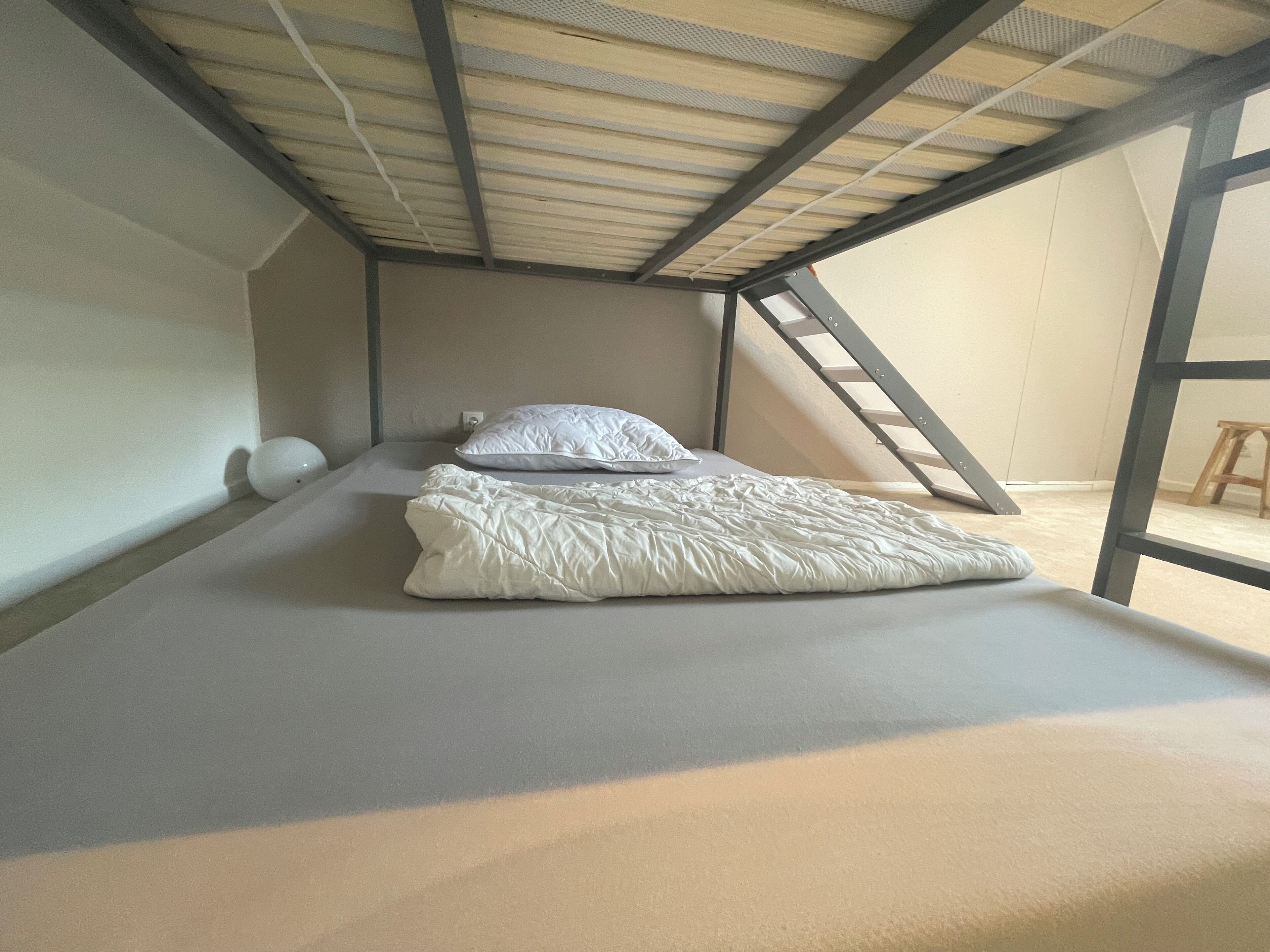 Een eenpersoonsbed met een witte dekbedovertrek in een kleine, afgesloten zolderkamer met een schuine plafond en een houten trap.