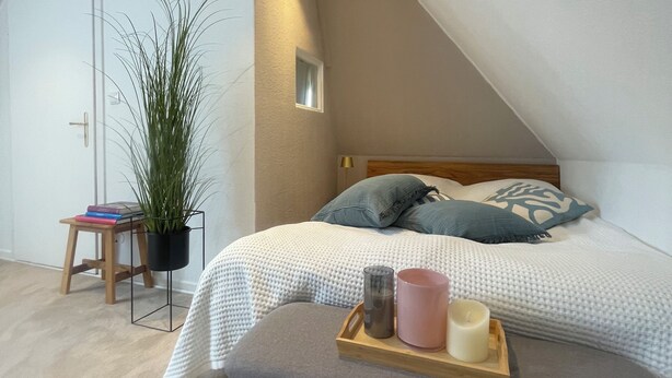 A modern bedroom with a sloped ceiling, a bed with a grey upholstered bench, a bedside table with books and a potted plant, and a small window. - Newly renovated vacation home - 4 min. to the beach - large, quiet lot surrounded by nature  (Schashagen)