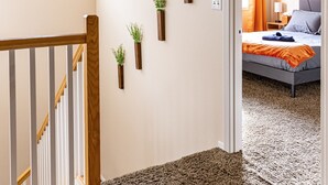 Ein Korridor mit Teppichboden, einer Holztreppe, einer Deckenleuchte und einer Tür zu einem Schlafzimmer mit Bett und Fenster.