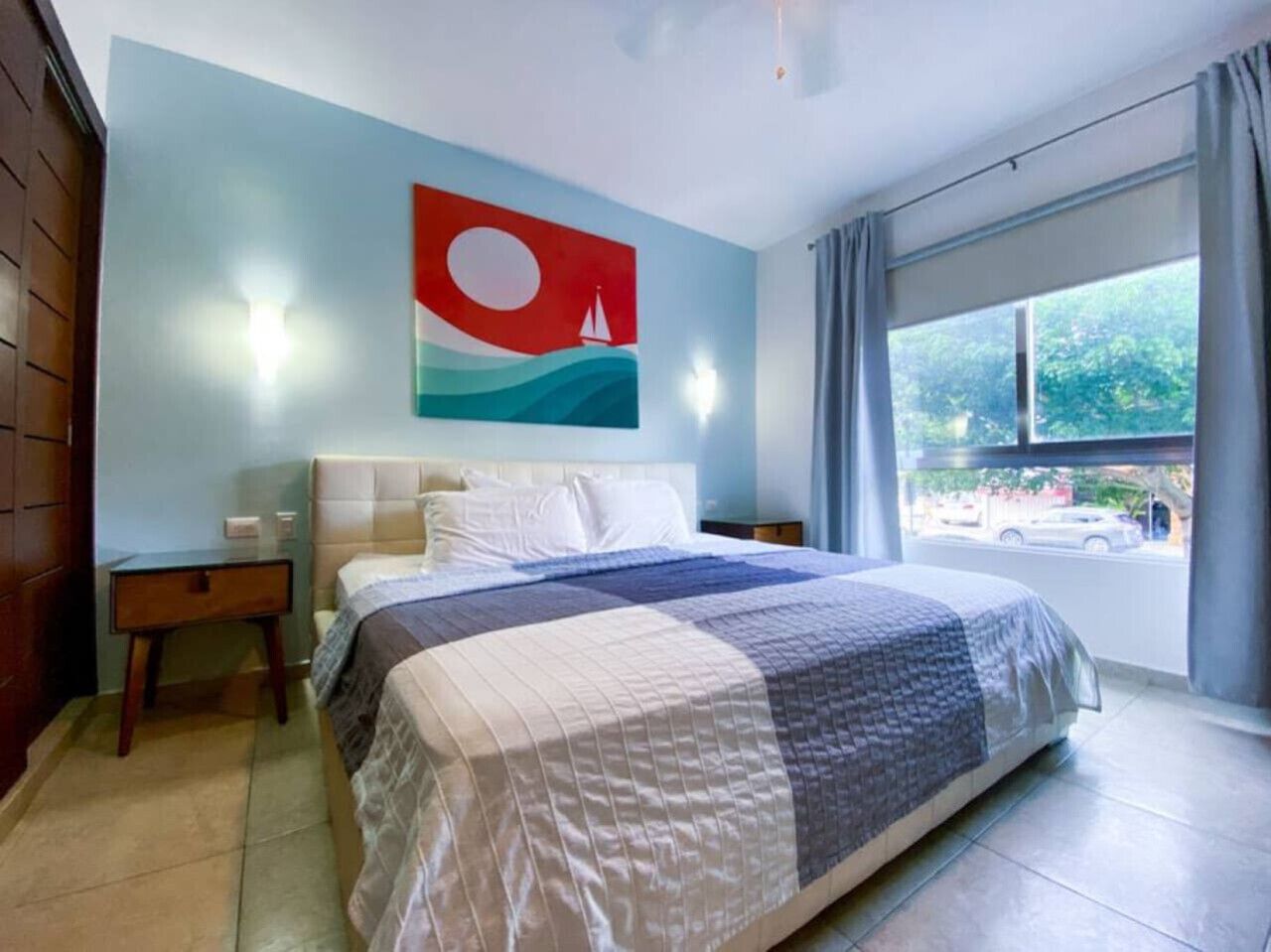 Une chambre à coucher comprenant un lit, une table de chevet, un bureau, une fenêtre avec des rideaux et un mur orné d’un tableau.
