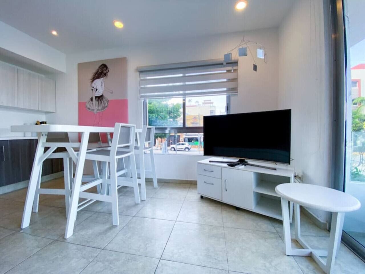 A modern living room with a flat-screen TV, white furniture, and a large window.