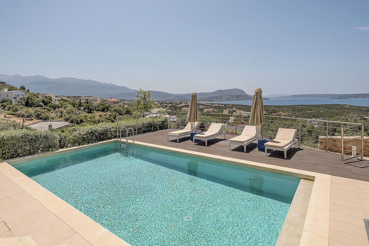 Ein Pool mit klarem Wasser, umgeben von Liegestühlen und Sonnenschirmen, mit Blick auf eine malerische Landschaft aus Bergen und einem Gewässer.