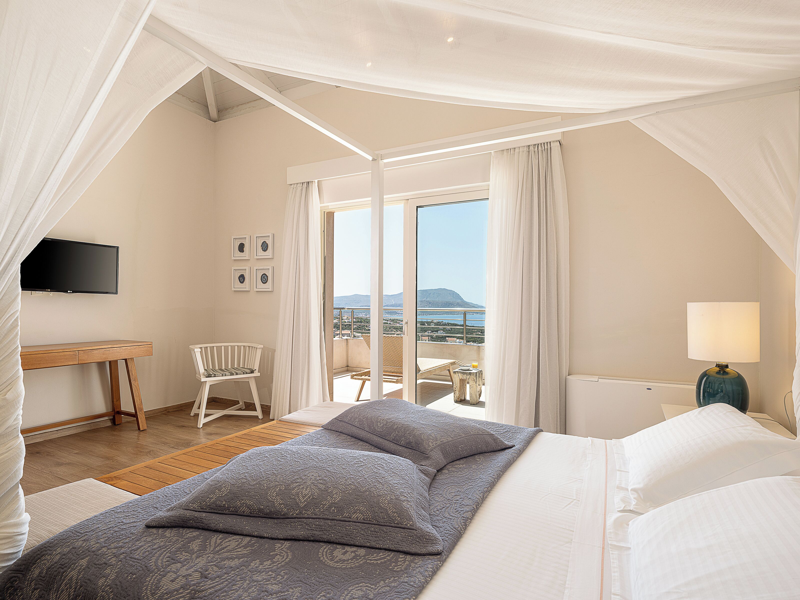 A bedroom with a bed, a desk, a chair, a TV, and a balcony with a view.