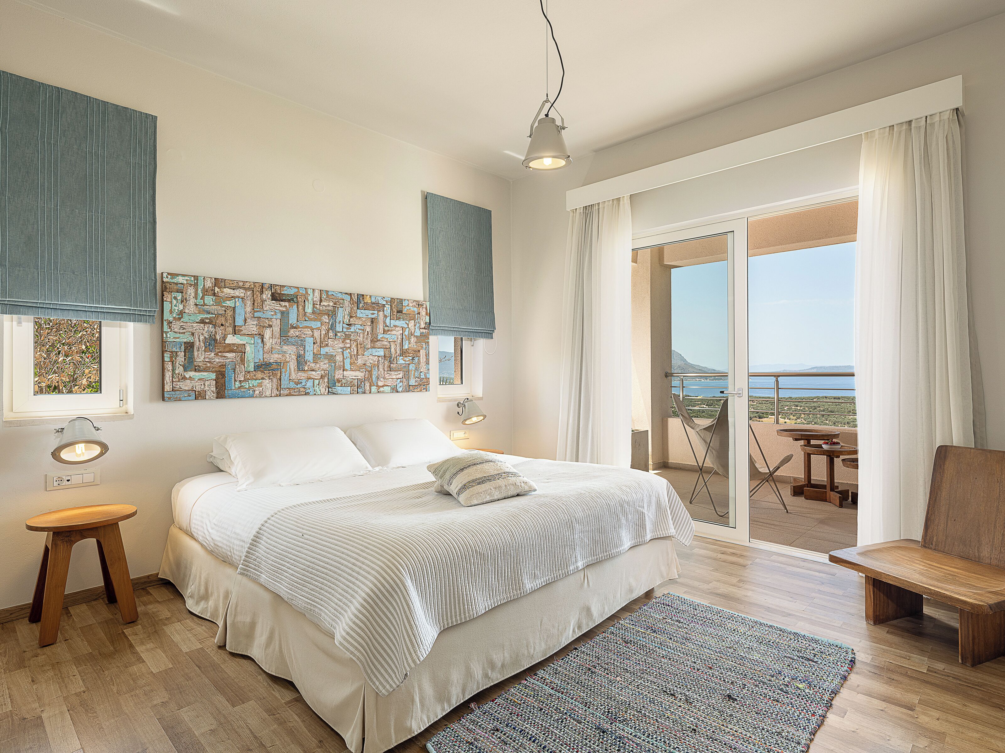 A bedroom with a bed, a nightstand, a lamp, a view of the balcony, and a large window.