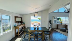 Een eetruimte met een tafel en stoelen, een houten bank, een spiegel en uitzicht op zee door een deur.