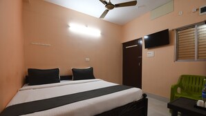 A bedroom with a bed, a ceiling fan, a TV, a window with blinds, and a green chair. - RADHA KRISHNA NIWAS (Puri)