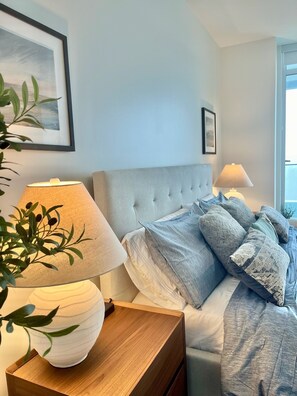 Ein ordentlich eingerichtetes Schlafzimmer mit einem gebürsteten Kopfende, einem Nachttisch mit Lampe, einer Zimmerpflanze und Blick ins Freie durch ein Fenster.