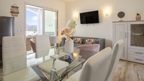 A modern living room with a glass dining table, white chairs, a grey sofa, and a TV mounted on the wall. - Victoria by Interhome (Budoni)