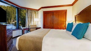Habitación de hotel con una cama grande, cabecera de madera, escritorio con lámpara y vistas a un bosque a través de amplios ventanales.