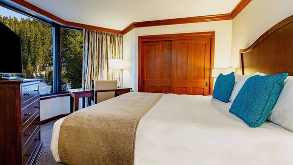 Ein Hotelzimmer mit einem großen Bett, einem Holzkopfende, einem Schreibtisch mit Lampe und Blick auf einen Wald durch große Fenster.