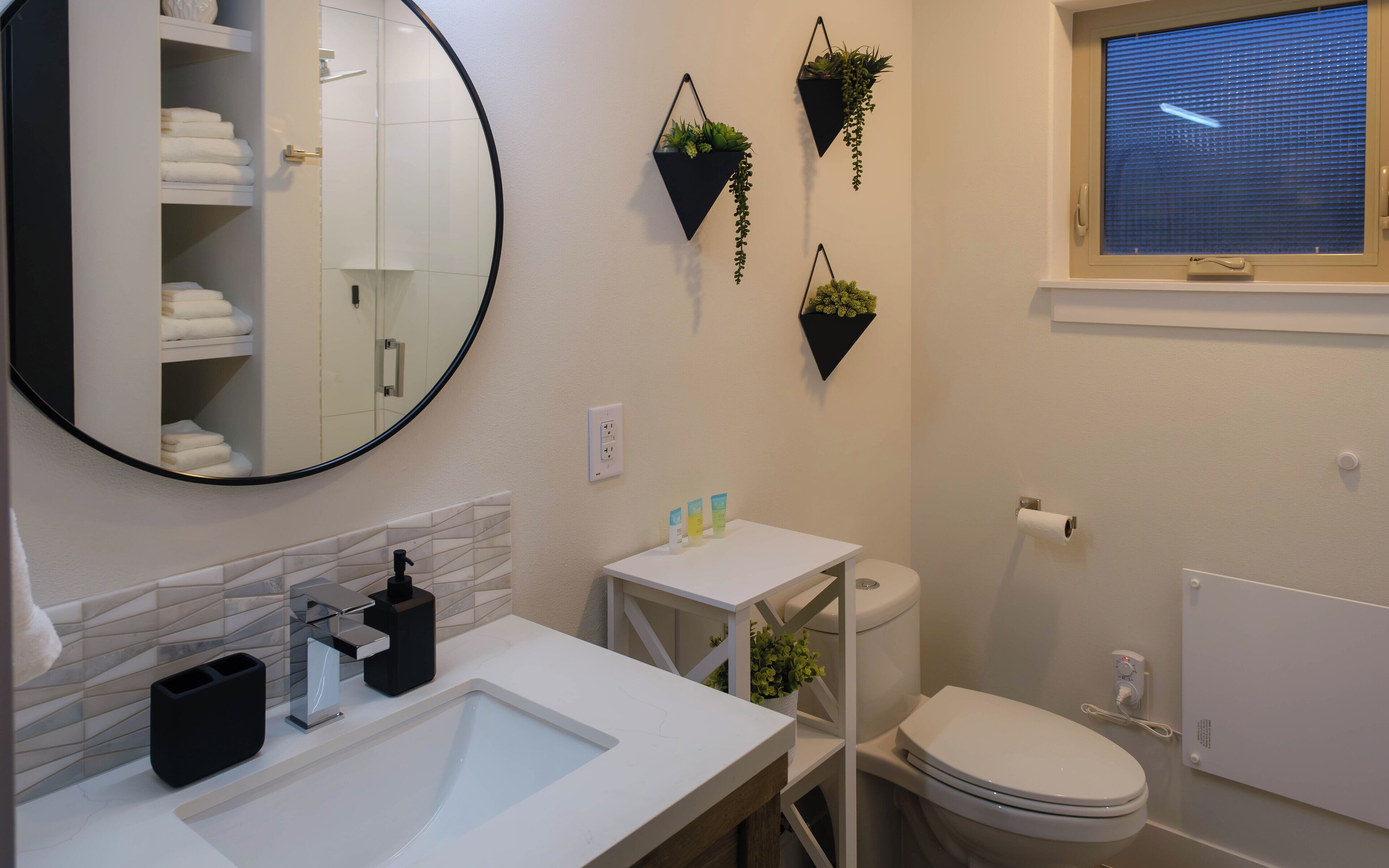 A modern bathroom with a round mirror, white sink, and a toilet. There are three hanging plants on the wall, and a window with blinds.