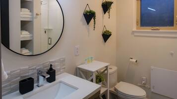 A modern bathroom with a round mirror, white sink, and a toilet. There are three hanging plants on the wall, and a window with blinds.