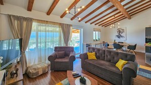 A spacious living room with a large sofa, a smaller armchair, and a television. The room features exposed wooden beams, a dining area, and a kitchen. - Vacation home Lynn by Interhome (Rogoznica)