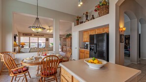 A spacious kitchen with wooden cabinets, a large island with a bowl of fruit, and a dining area with wooden chairs and a table. - Covered Patio, Lava Rock Gas Fire Pit, Near Spring Training, Golf, Hiking & Biking; 30 Day Min (Gilbert)