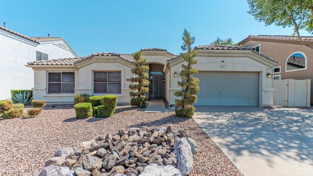 A residential house with a garage, a driveway, and a landscaped front yard with rocks and trimmed bushes. - Covered Patio, Lava Rock Gas Fire Pit, Near Spring Training, Golf, Hiking & Biking; 30 Day Min (Gilbert)