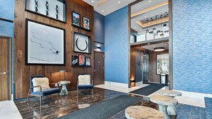 A modern lobby with a wooden wall, framed artwork, a marble floor, and a seating area with a fluffy chair and a small table.