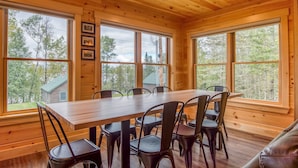 A wooden interior with a dining table and chairs, large windows, and framed pictures on the wall.