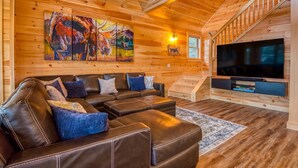 A cozy living room with a sectional sofa, a flat-screen TV, wooden walls, and a staircase.