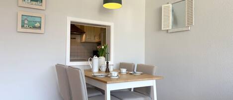 A dining area with a table, chairs, a hanging light fixture, and a kitchen visible through an open window.