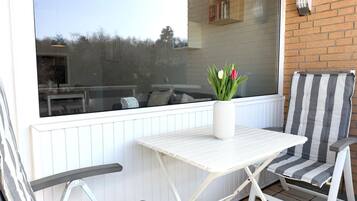 Une terrasse avec une table et des chaises blanches, une fenêtre donnant sur un mur de briques et une bibliothèque, ainsi qu’un vase rempli de fleurs.