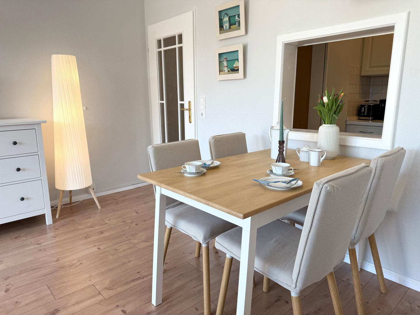 A dining area with a wooden table, four chairs, a lamp, and a view of the kitchen through a window.