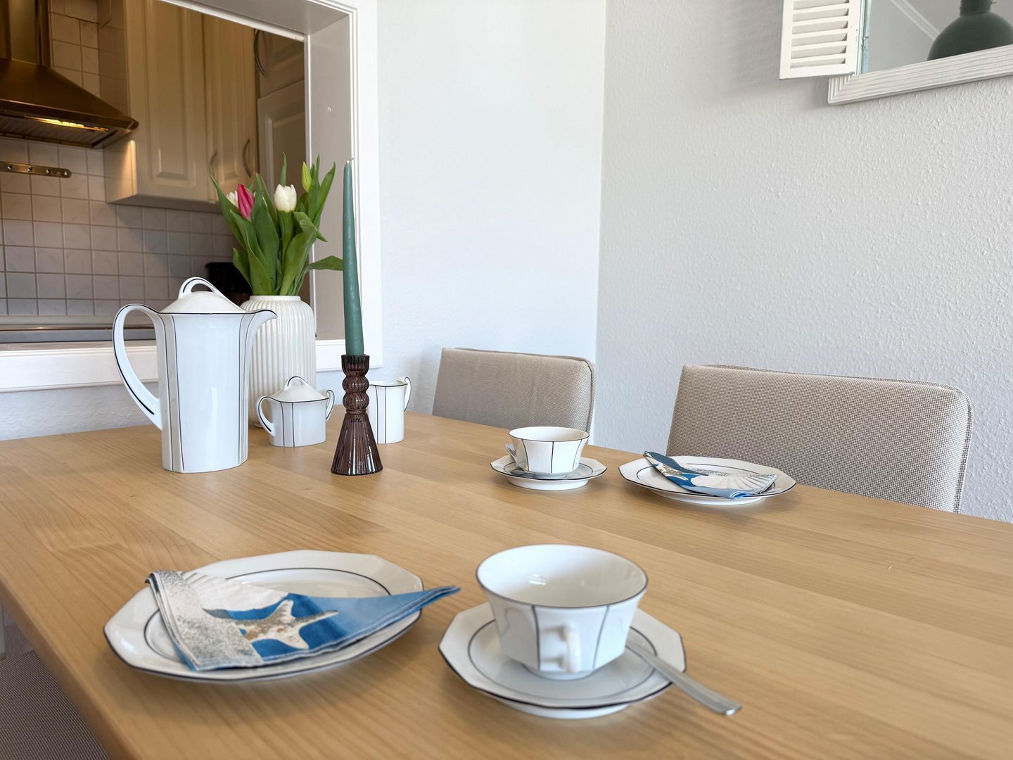 Un comedor con una mesa de madera dispuesta para dos personas, vajilla blanca y un jarrón con flores.