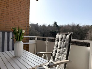 Een balkon met een witte tafel, een stoel met een geruit kussen en een vaas met bloemen.