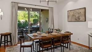 A dining area with a wooden table set for four, surrounded by chairs, with a view of greenery outside through large glass doors. - Luxe 4-BR Home: Spa, Red Rock Views, and A+ Trails (Sedona)