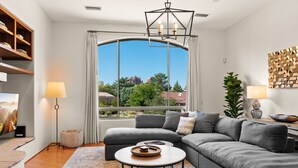 A modern living room with a grey sectional sofa, a round coffee table, and a large window offering a view of greenery and buildings. - Luxe 4-BR Home: Spa, Red Rock Views, and A+ Trails (Sedona)