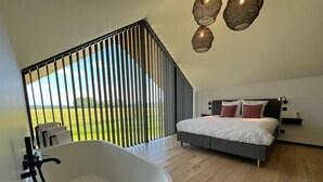 A bedroom with a large bed, a bathtub, and a view of a green field through a window with vertical blinds. - Lodge in Durbuy With Hot Tub & Valley Views (Durbuy)