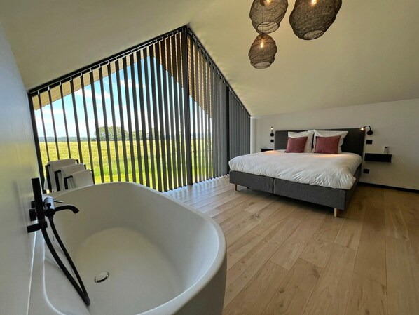 A bedroom with a large bed, a bathtub, and a view of a green field through a window with vertical blinds. - Lodge in Durbuy With Hot Tub & Valley Views (Durbuy)