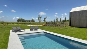A rectangular swimming pool with clear blue water, surrounded by a paved deck. There are four lounge chairs aligned along the pool's edge. The pool is set against a backdrop of a grassy lawn, a wooden fence, and a large building to the right. - Lodge in Durbuy With Hot Tub & Valley Views (Durbuy)
