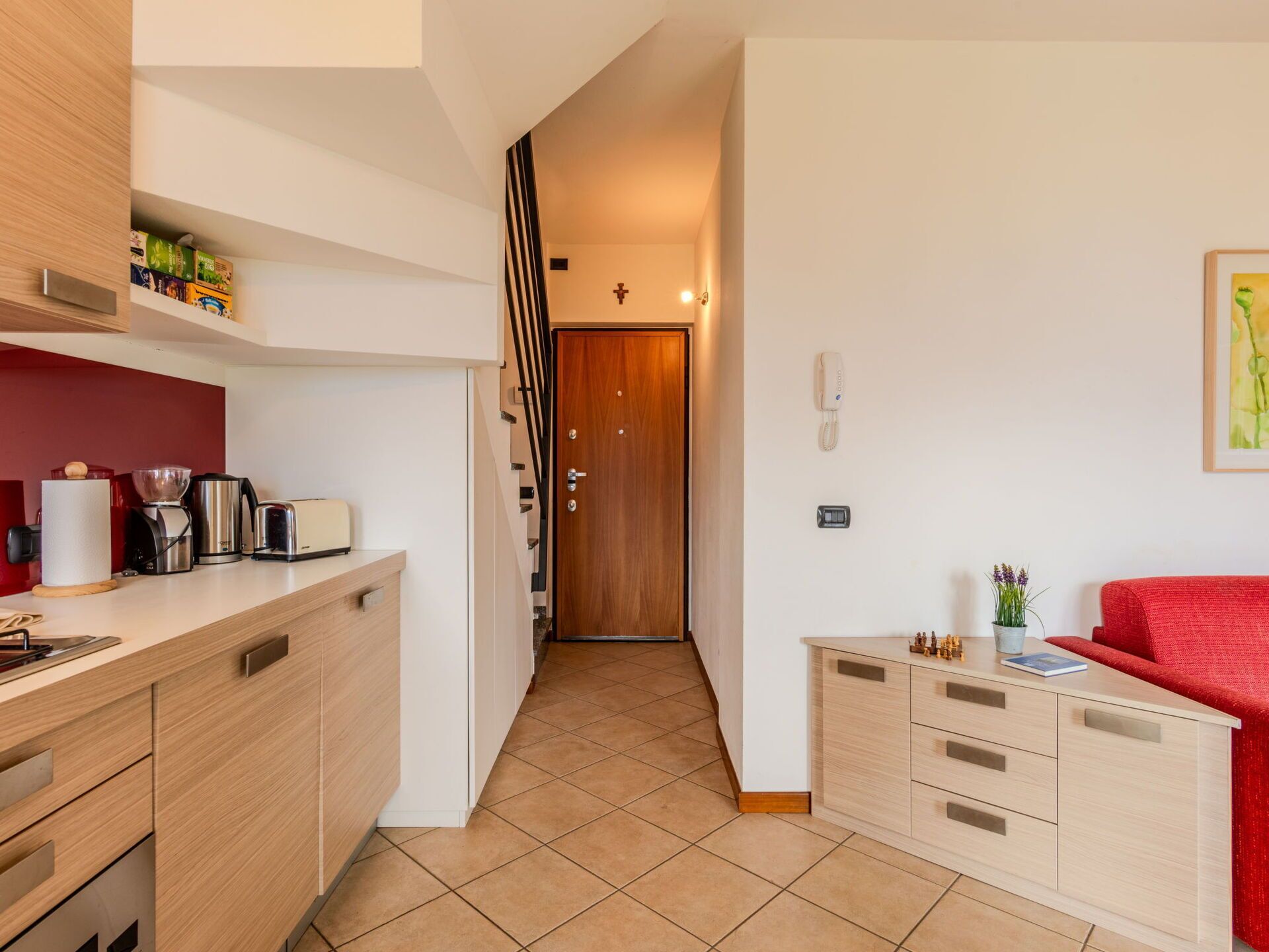 A modern kitchen with a coffee maker, toaster, and cabinets. A wooden door leads to a hallway with a cross on the wall.