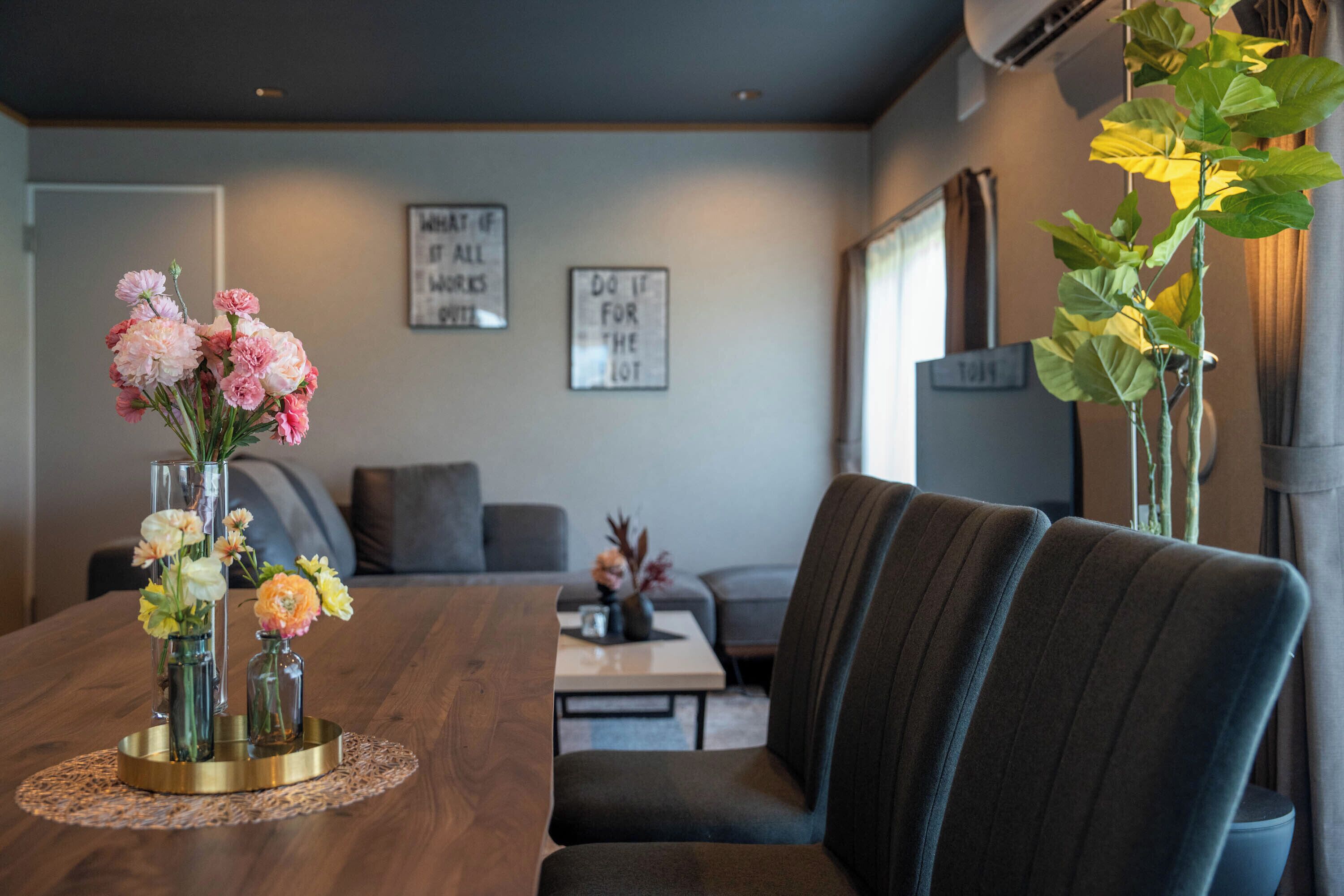Un comedor con una mesa de madera, sillas y un florero con flores. Hay dos letreros enmarcados en la pared y una planta a la derecha.