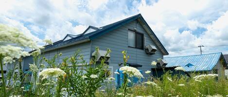 Una casa con techo inclinado y fachada azul, rodeada de exuberante follaje y flores blancas.